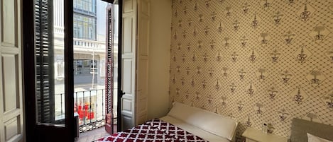 Un dormitorio con cama, mesita de noche, ventana con contraventanas y un papel tapiz estampado.
