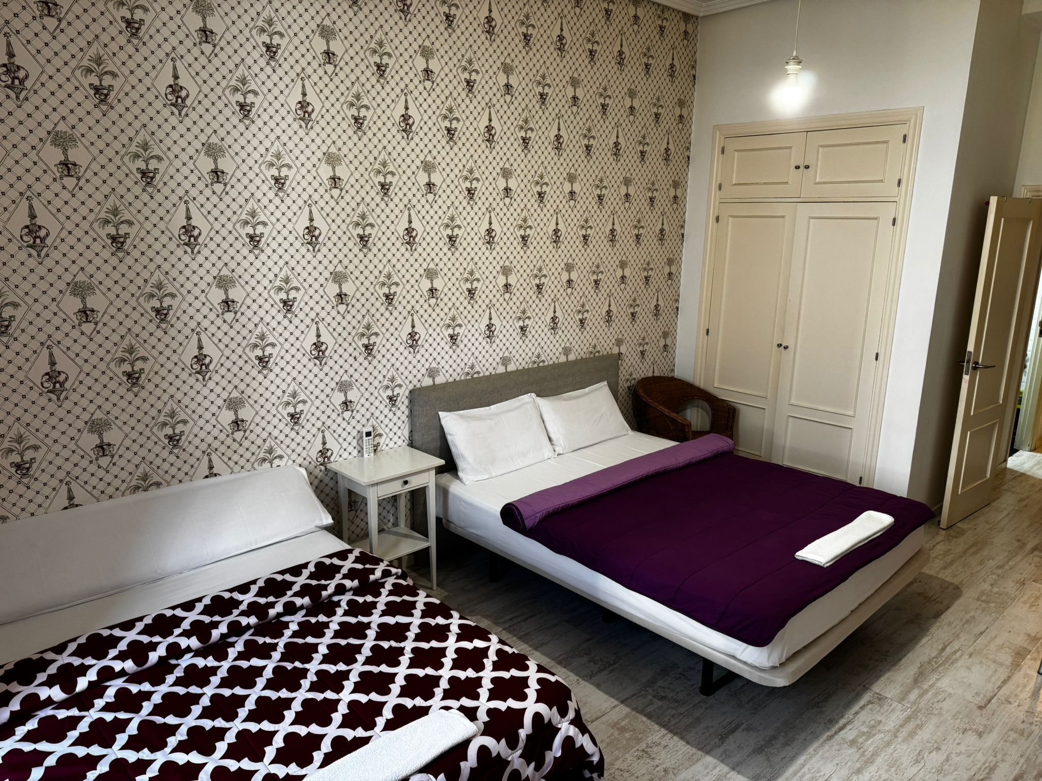 A bedroom with a bed, a bedside table, a wardrobe, and a patterned wallpaper.