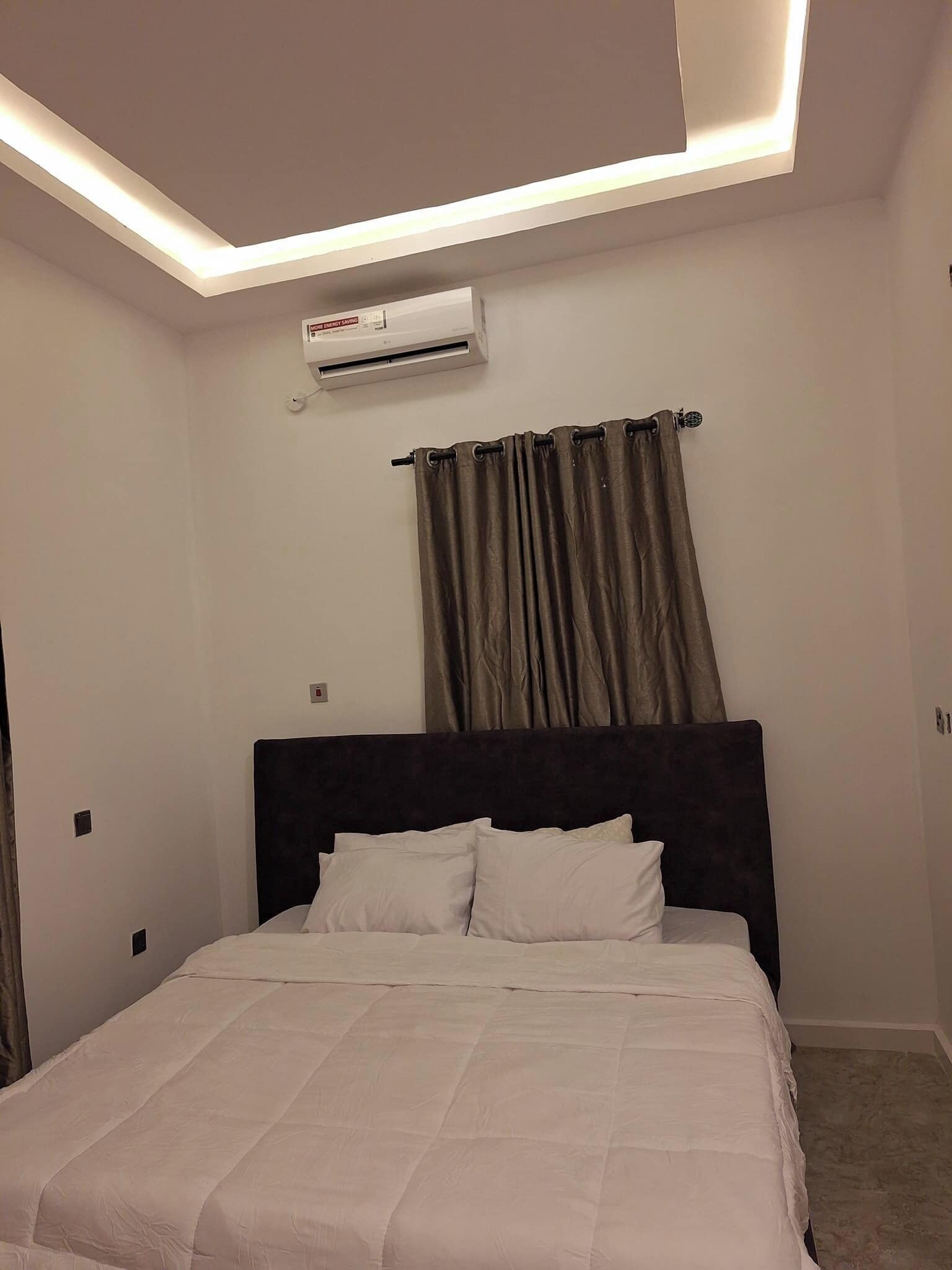 Um quarto com cama, cabeceira, janela com cortinas e ar condicionado.