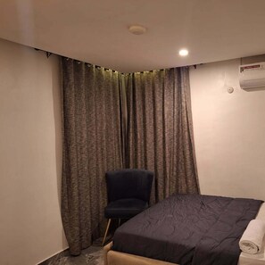 A small, neatly arranged bedroom with a bed, a chair, and a window with curtains. - The Royals Apartment  (Ibadan)