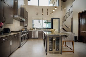 A modern kitchen with stainless steel appliances, a central island, and a dining area with a sofa. - Ocean View Three Bedroom Villa (3 Kings) - Prana Maya Island Resort (Placencia)
