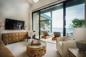 A modern living room with a large TV, comfortable seating, a glass coffee table, and a view of the sea. - Ocean View Three Bedroom Villa (3 Kings) - Prana Maya Island Resort (Placencia)
