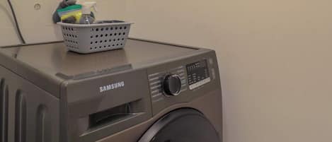 A Samsung washing machine in a laundry room with a basket of laundry on top.