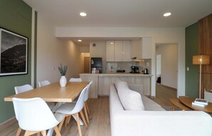 Uma cozinha moderna com armários brancos, uma sala de jantar com mesa de madeira e cadeiras brancas, e uma sala de estar com sofá branco.