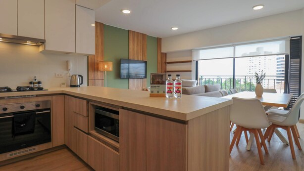 A modern kitchen with a central island, a dining area with a table and chairs, a flat-screen TV mounted on the wall, and a large window offering a view of the city. - w* | Welcoming 2BR w/ Balcony in Miraflores (Miraflores)