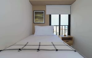 A single bed with a checkered bedspread, a white pillow, and a nightstand with a lamp. A window with a view of buildings is visible. - w* | Upscale 3BR w/ Balcony in Upscale Miraflores (Miraflores)