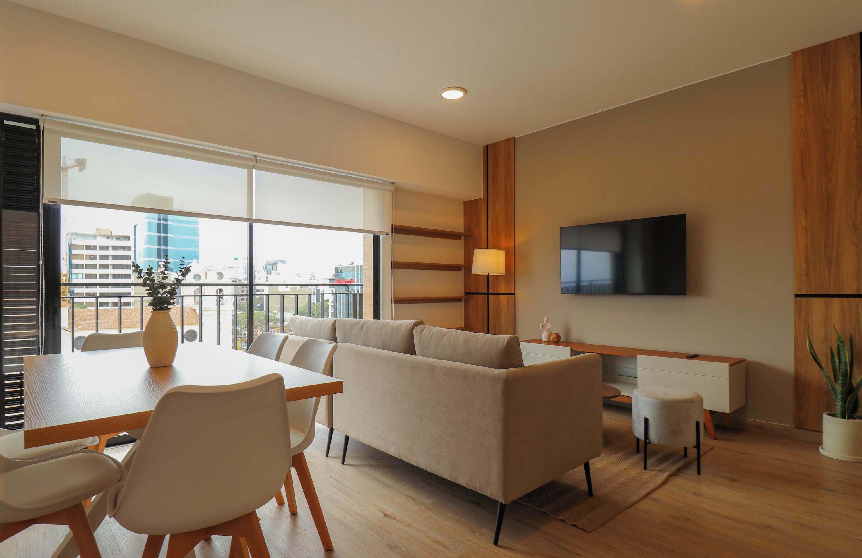 A modern living room with a sofa, dining table, chairs, a TV, and a balcony with a city view.