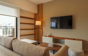 A modern hotel room with a large window, a flat-screen TV mounted on the wall, a wooden accent wall, a sofa, a coffee table, and a small side table with a lamp. - w* |  Airy 2BR w/ Balcony in Prime Miraflores (Miraflores)
