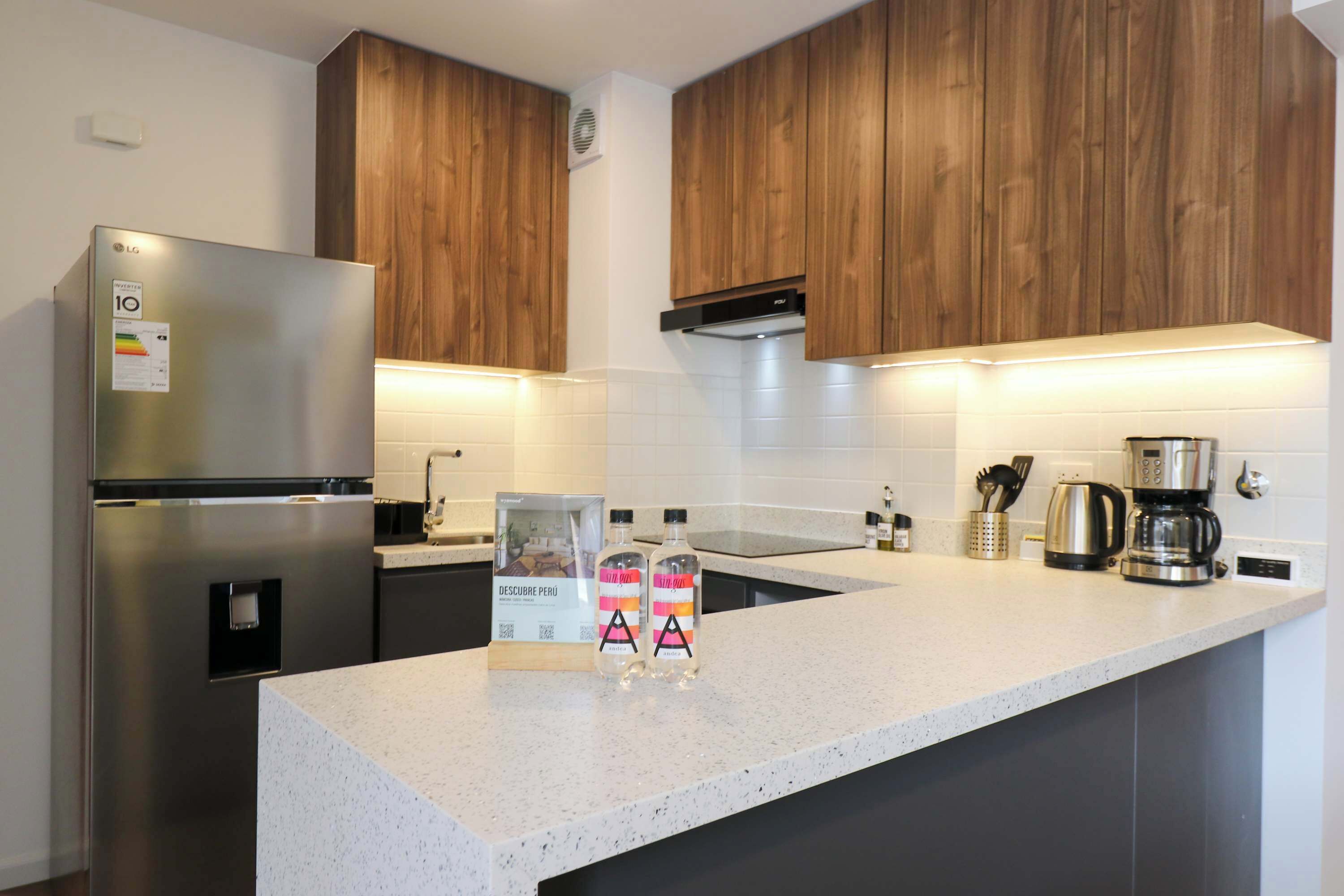 A modern kitchen with wooden cabinets, a stainless steel refrigerator, a coffee maker, and two bottles of a beverage.