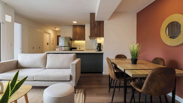 A modern living room with a sofa, a dining table with chairs, and a kitchen area in the background. - w* | Prime 2BR w/ Balcony in Miraflores (Miraflores)