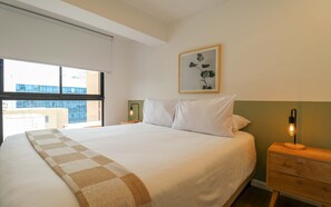 A hotel room with a large bed, a bedside table with a lamp, a window with a view of buildings, and a framed picture on the wall. - w* | Prime 2BR w/ Balcony in Miraflores (Miraflores)