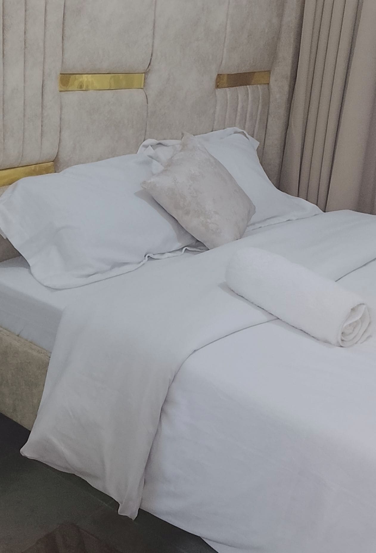 A neatly made bed with white linens and pillows, a rolled towel, and a headboard with a gold accent.