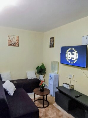 A living room with a sofa, a small round table with a vase, a water dispenser, and a flat-screen TV mounted on the wall. - Budget studio apartment in charming Rungiri Waiyaki way perfect for relaxing (Rungiri)