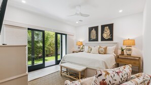 Ein Schlafzimmer mit einem großen Bett, zwei Nachttischlampen, einem Flachbildfernseher, einer Bank und Blick auf Grün hinter einer Glasschiebetür.