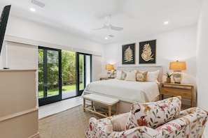 A bedroom with a large bed, two bedside lamps, a flat-screen TV, a bench, and a view of greenery through the sliding glass door. - Villa Del Mar - East Beach (St Simons Island)