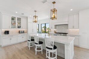 Uma cozinha moderna com armários brancos, uma ilha central com banquetas, iluminação pendente e vista para o exterior.