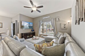 Ein Wohnzimmer mit Deckenventilator, einem großen Fenster mit Jalousien, einem Flachbildfernseher und einer Couch mit Zierkissen.
