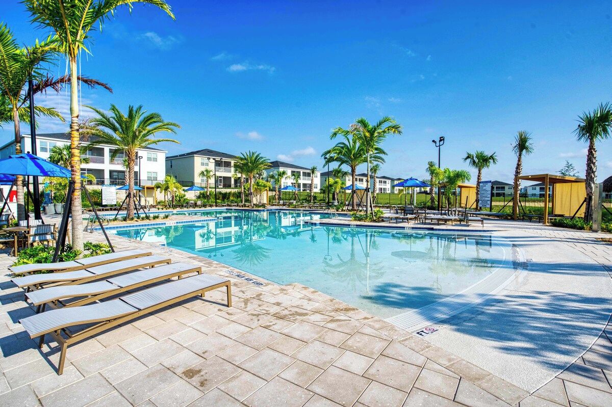A swimming pool with lounge chairs and palm trees, surrounded by buildings.