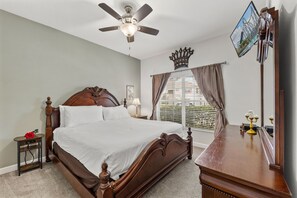 A bedroom with a wooden bed, a nightstand, a bedside table, a ceiling fan, a window with blinds, a mirror, and a wall-mounted TV.