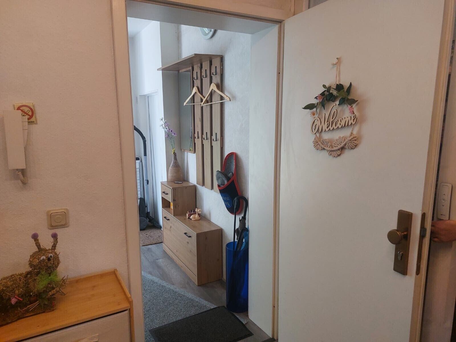 Een hal met een houten kast, een vaas met bloemen, een bord met de tekst welkom en een kapstok met ophanghaakjes.