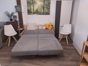 Un dormitori modern amb un sofà llit gris, una petita taula de nit amb una làmpada, un quadre al mur i una planta en test.