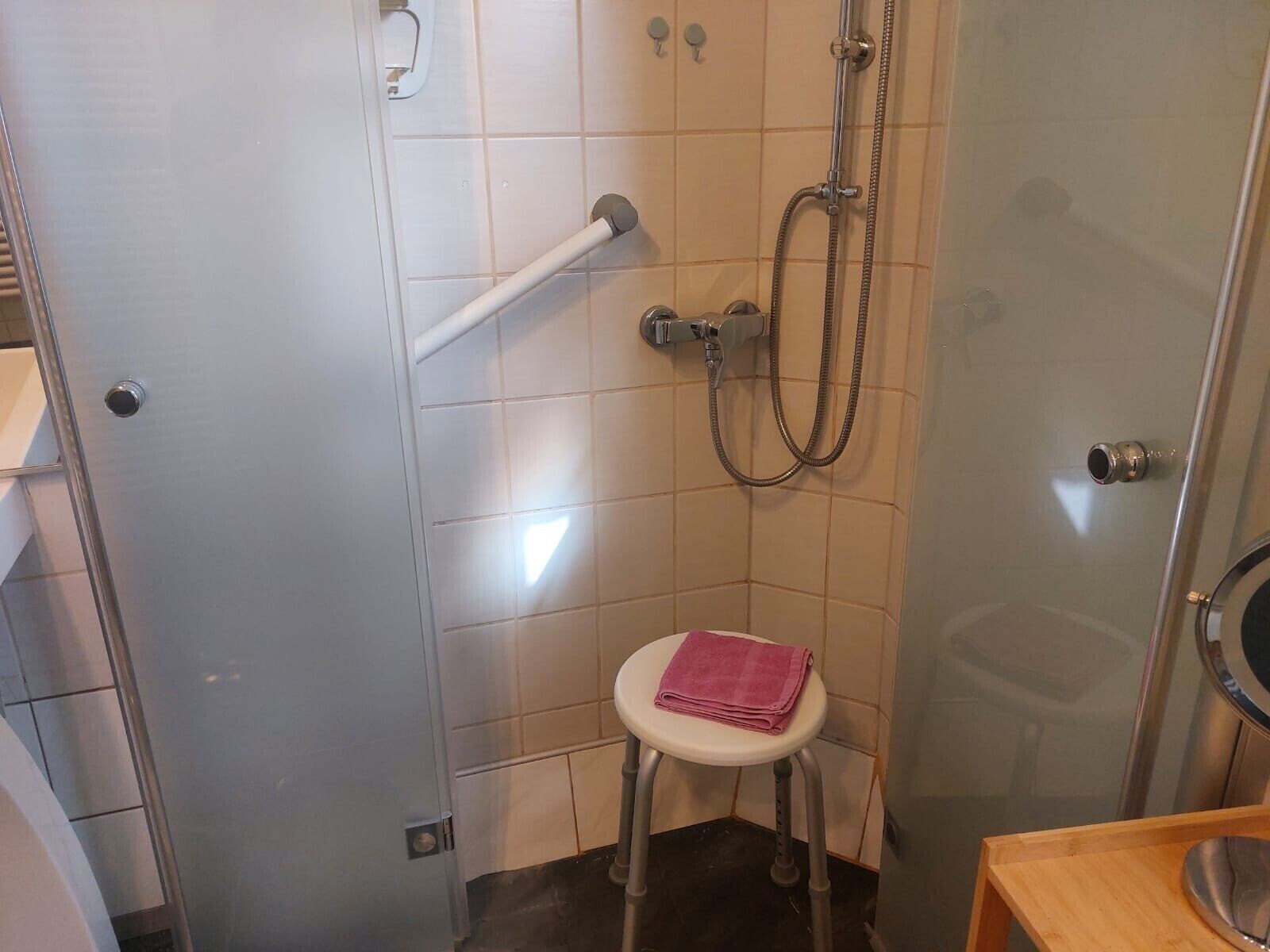 A bathroom with a glass shower enclosure, a white towel on a stool, and a wooden vanity.