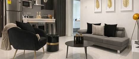 A modern living room with a sofa, armchair, coffee table, and a kitchen area with appliances.
