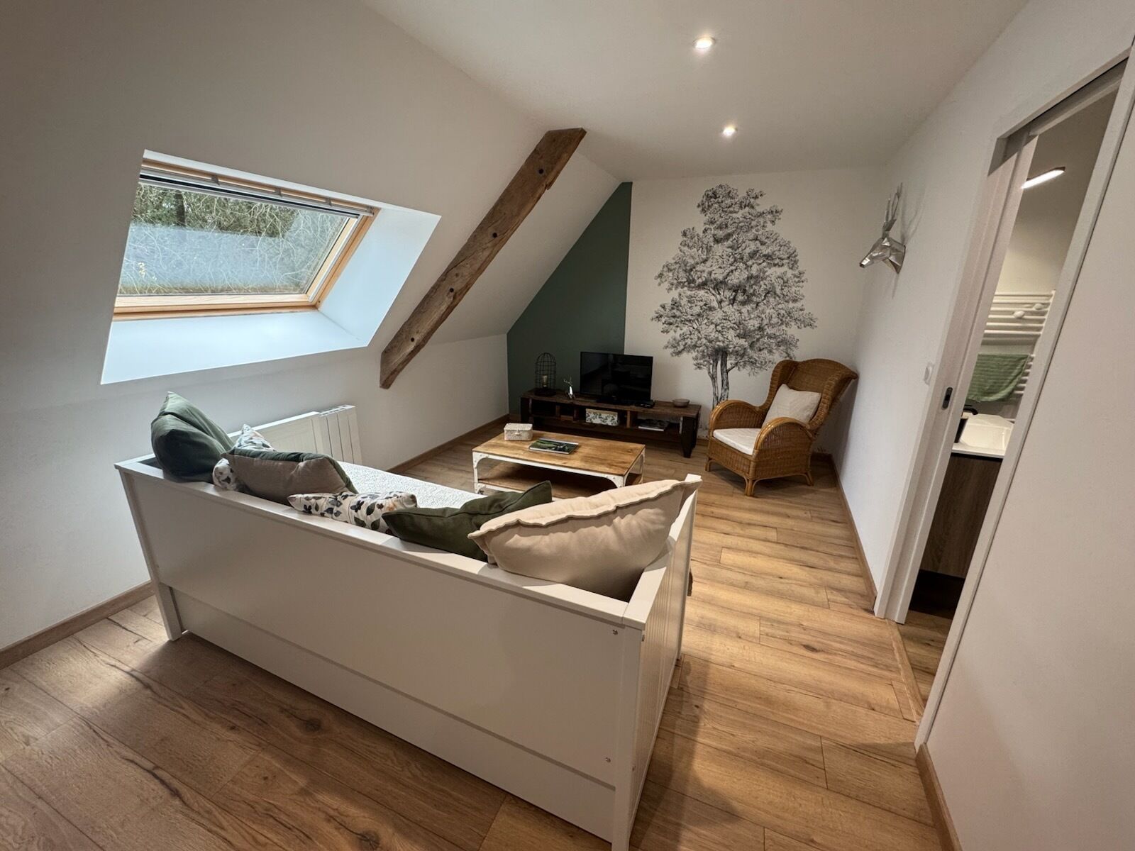 Un salón estilo loft con techo inclinado, televisor empotrado, sofá blanco, sillón de mimbre y un ventanal grande.