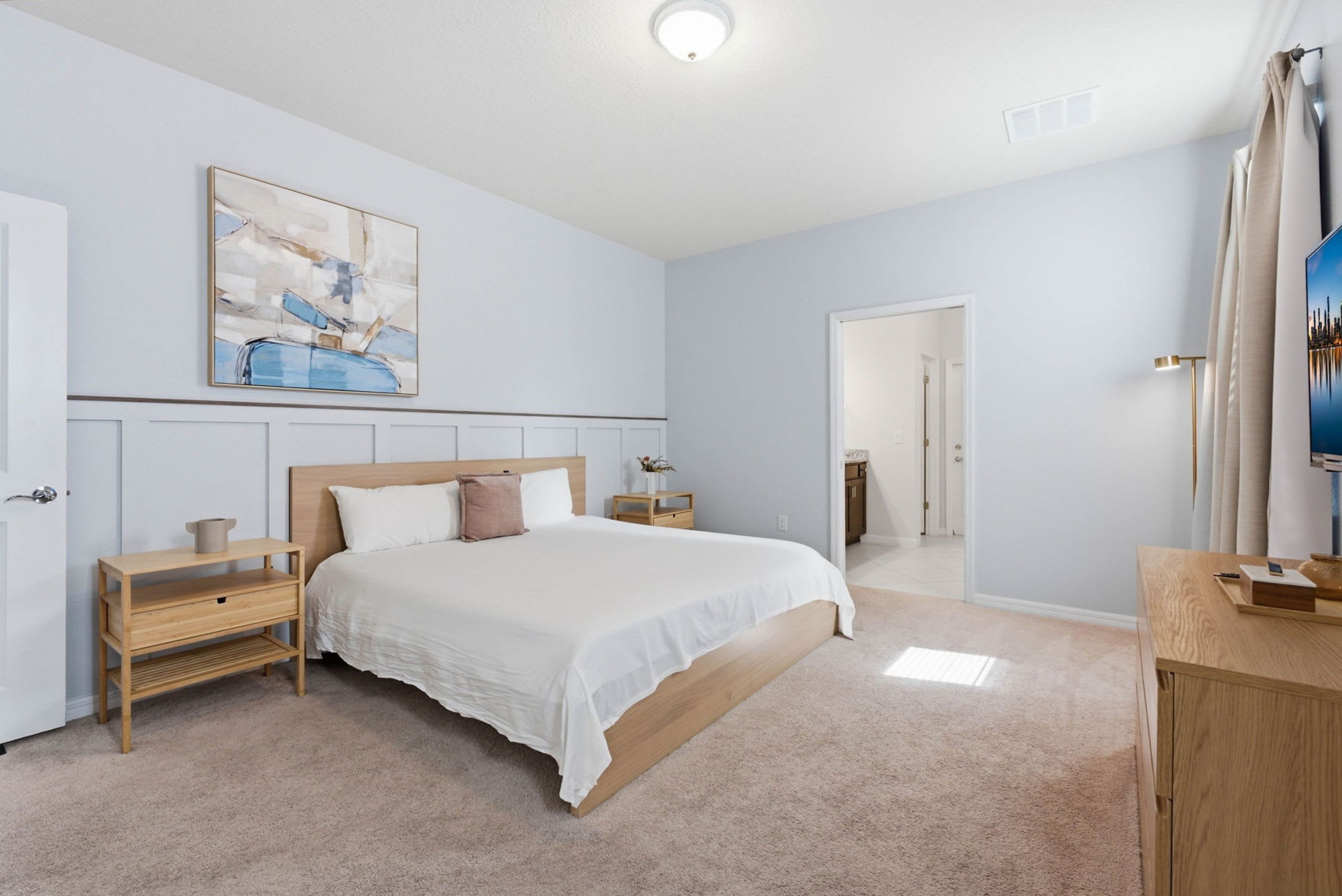 A bedroom with a bed, bedside tables, a nightstand, a painting, a mirror, and a door leading to another room.