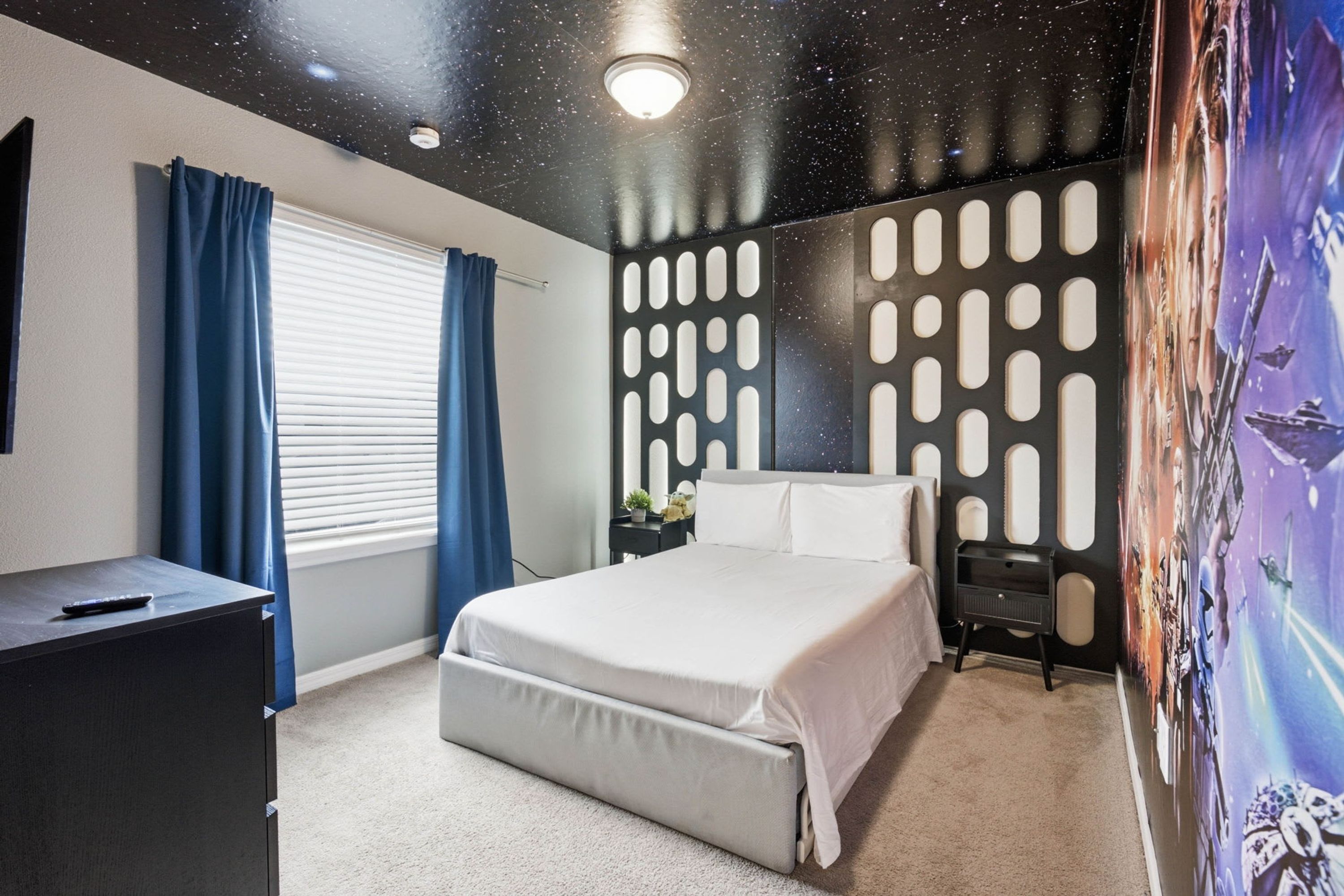 A bedroom with a bed, a desk, a dresser, a window with curtains, and a wall with a unique pattern.