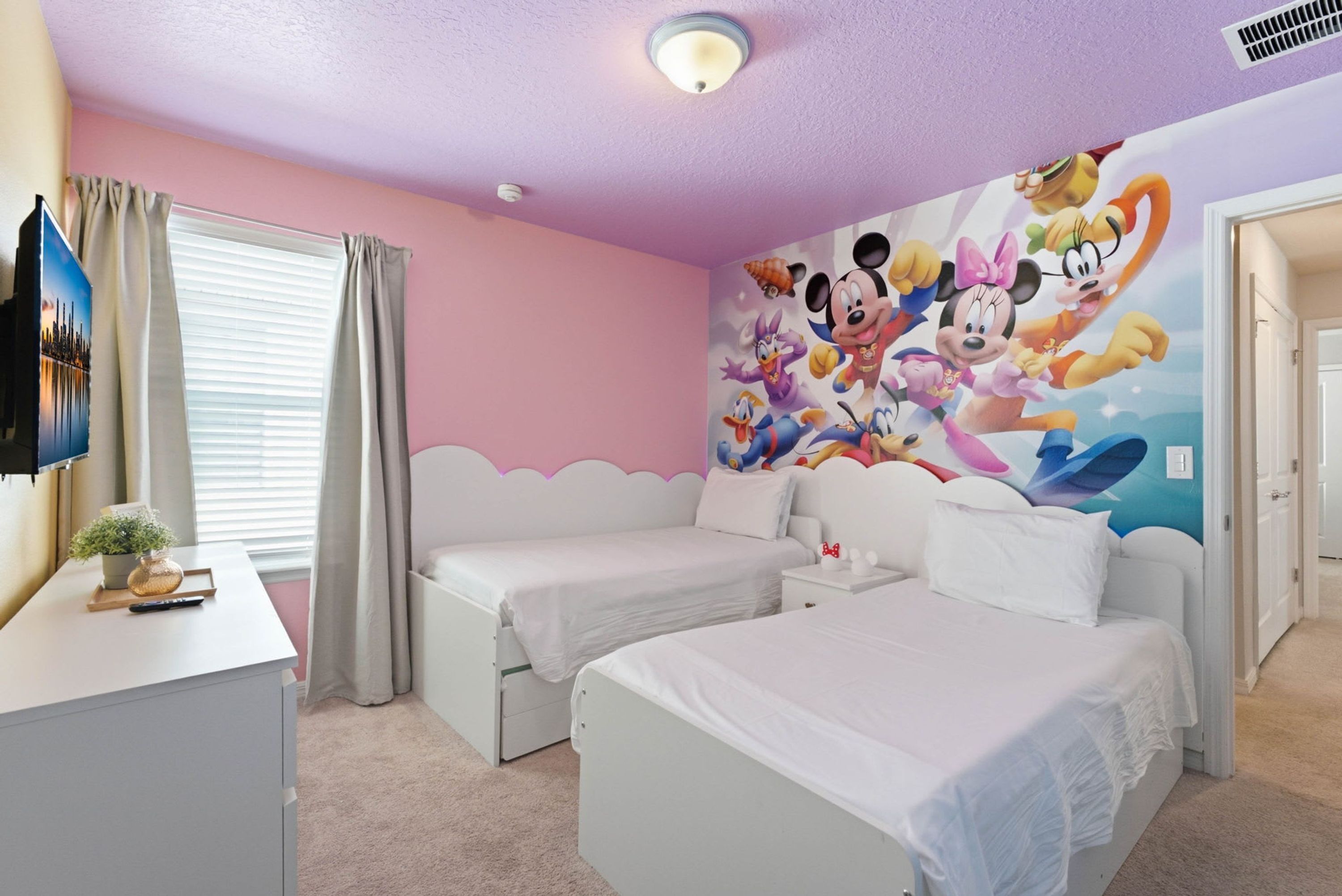 A bedroom with two single beds, a flat-screen TV, a decorative wall mural, and a window with curtains.