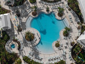Luftaufnahme eines Resorts mit einem zentralen Schwimmbecken, umgeben von Liegebereichen mit Stühlen und Tischen sowie Palmen.