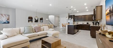 A modern living room with a sectional sofa, a coffee table, and a kitchen area with dark cabinets and a large island.
