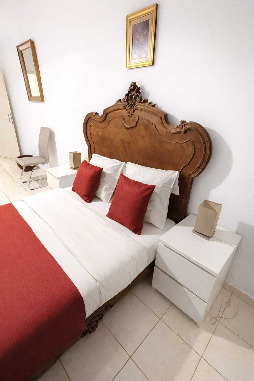 Een hotelkamer met een houten hoofdeinde, een bed met wit en rode beddengoed, een nachtkastje met een lamp en een klein nachtkastje met een doos.