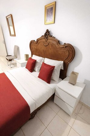 A hotel room with a wooden headboard, a bed with white and red bedding, a bedside table with a lamp, and a small bedside table with a box.