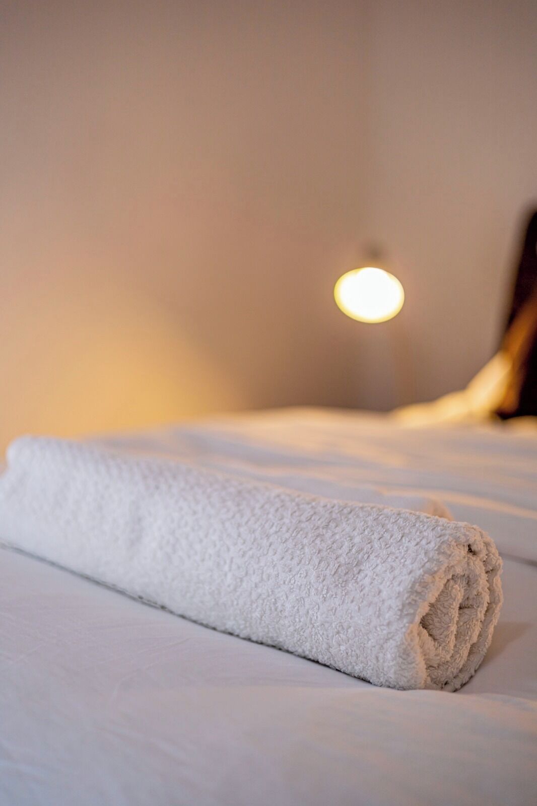 Un asciugamano bianco piegato con cura su un letto, con una lampada a parete sullo sfondo.