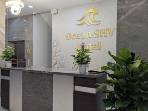 Un mostrador de recepción con un cartel que dice Ocean SHV Hotel, un teléfono y plantas en macetas.