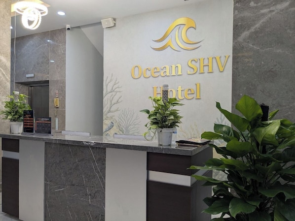Un comptoir d’accueil avec une enseigne indiquant « Ocean SHV Hotel », un téléphone et des plantes en pot.