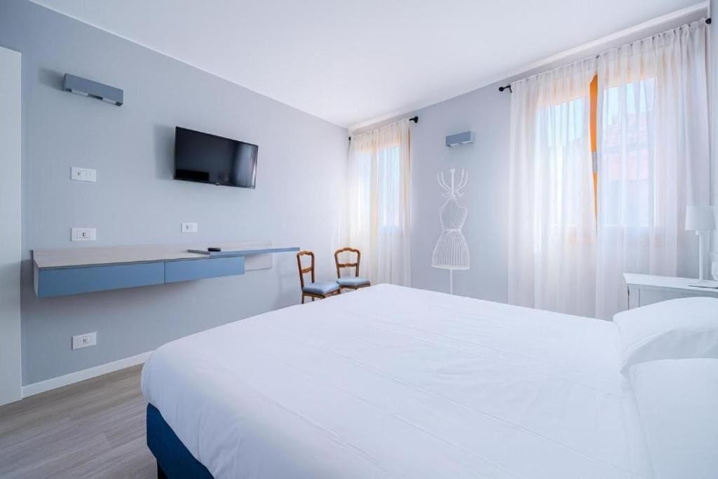 A modern hotel room with a large bed, a flat-screen TV mounted on the wall, a desk with a chair, and a window with sheer curtains.