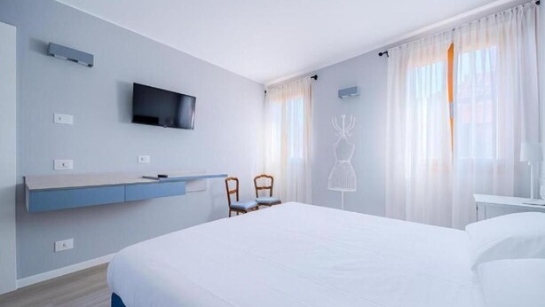 Ett modernt hotellrum med en stor säng, en platt-TV som är monterad på väggen, ett skrivbord med en stol och ett fönster med tunna, genomskinliga gardiner.