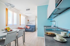 A modern kitchen with a blue accent wall, a dining area with a table and chairs, and a living room with a sofa and a vase of flowers. - Monet Suite, 100 meters from the Biennale - Canal View (Venezia)