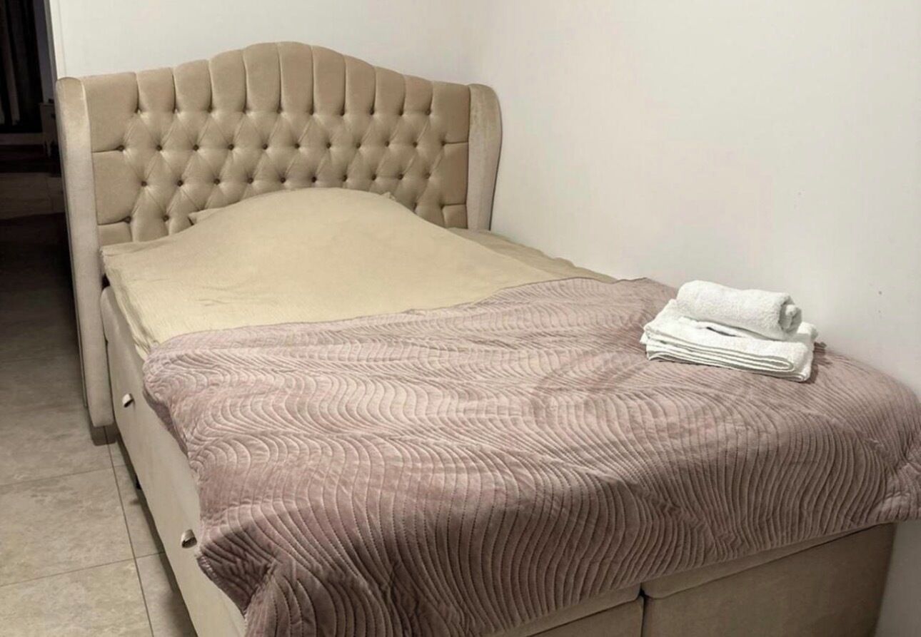 Een bed met een gestoffeerd hoofdbord, een beige matras en een opgevouwen witte handdoek aan het voeteneinde.