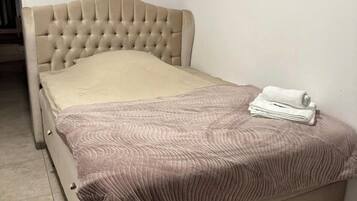 Een bed met een gestoffeerd hoofdbord, een beige matras en een opgevouwen witte handdoek aan het voeteneinde.