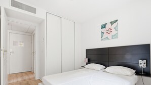 Un dormitori amb un llit, una tauleta de nit, una làmpada penjada a la paret i una porta que dóna a una altra habitació.