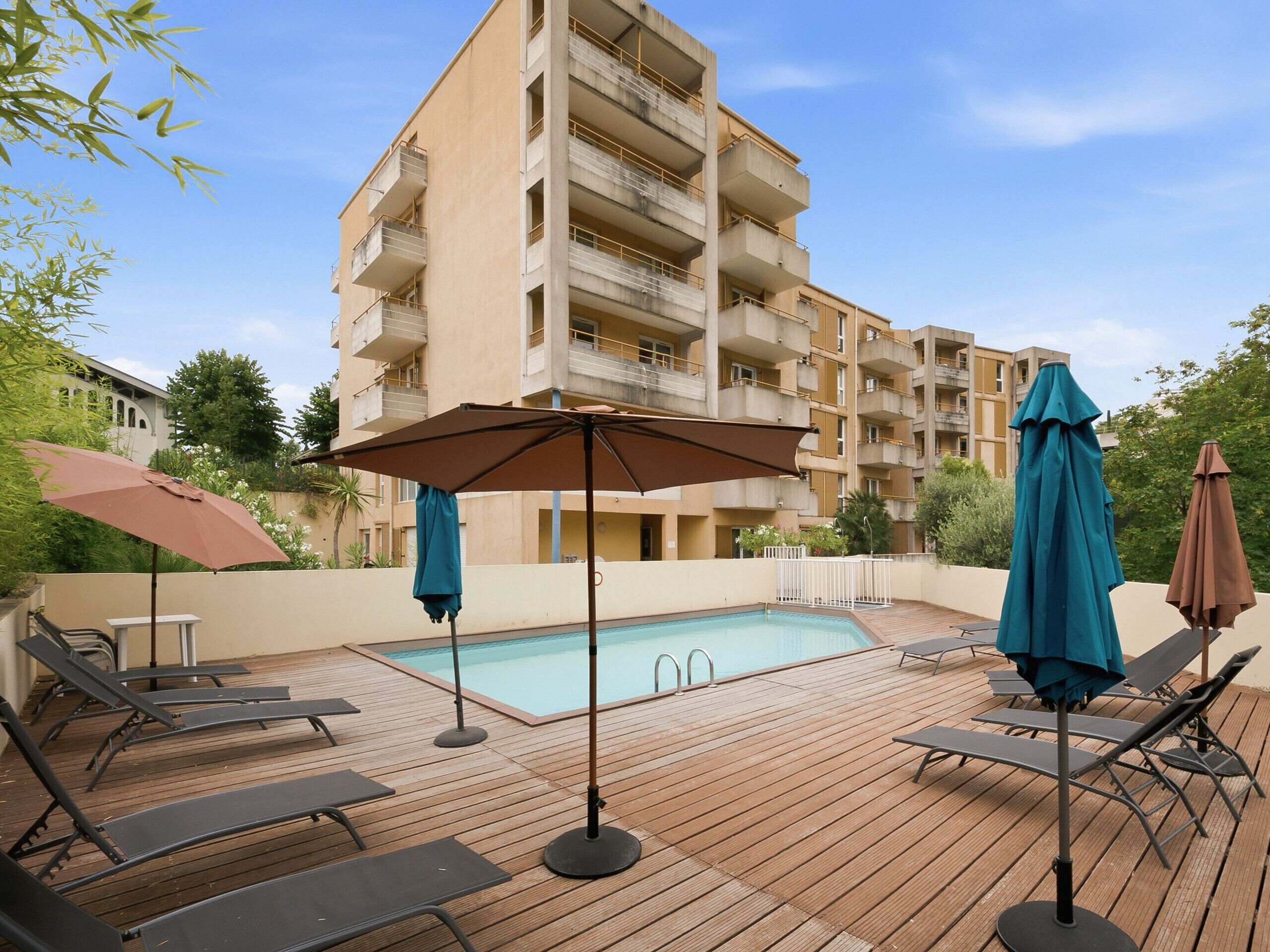 A poolside area with lounge chairs and umbrellas, a pool, and a multi-story building in the background.