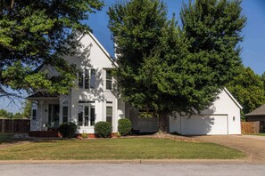 A two-story white house with a front porch, a garage, and a large tree in the yard. - Bentonville Belle • Bike Storage & Trails (Bentonville)