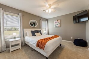 A bedroom with a bed, a nightstand, a mirror, a ceiling fan, a window with blinds, a painting, and a TV mounted on the wall. - Bentonville Belle • Bike Storage & Trails (Bentonville)