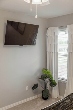 A room with a flat-screen TV mounted on the wall, a window with white curtains, and a potted plant. - Bentonville Belle • Bike Storage & Trails (Bentonville)