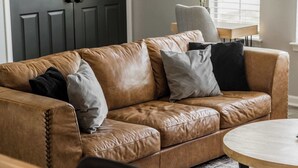 Ein braunes Ledersofa mit mehreren Kissen, ein runder Holztisch und eine graue Tür im Hintergrund.