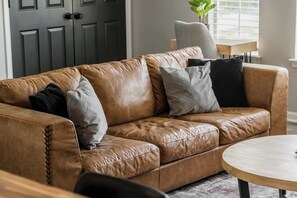A brown leather sofa with multiple cushions, a round wooden coffee table, and a grey door in the background. - Double Black Diamond | Trails | Bike Out | 2 Decks (Bentonville)
